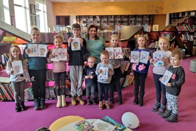 Grupa dzieci z opiekunem i laurkach w rękach stoją i pozują na tle czytelni dziecięcej.