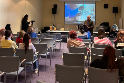 Sala konferencyjna pełna ludzi. Józef Lewkowicz - w tle z drzeworytem w ręckach i filmie o drzeworycie w tle.