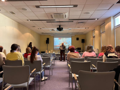 Sala konferencyjna pełna ludzi. Józef Lewkowicz - w tle z drzeworytem w ręckach i filmie o drzeworycie w tle.