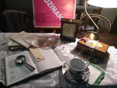 Kompozycja - Na stole znajduję się mnóstwo elementów. Na samym środku znajduję się ulotka Klubu Dyskusyjnego Książka, która jest otoczona równymi elementami. Na dole filiżanka z talerzykiem na haftowanej chusteczce, wyżej szkatułka z biżuterią. Nad ulotką zegar, a po lewej stronie książki i kolejna chusteczka. Po lewej stronie od ulotki mamy otwartą książka z lupą i zakładką. W tle znajduje się różowy baner z białym napisem o klubie.