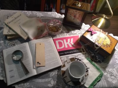 Kompozycja - Na stole znajduję się mnóstwo elementów. Na samym środku znajduję się ulotka Klubu Dyskusyjnego Książka, która jest otoczona równymi elementami. Na dole filiżanka z talerzykiem na haftowanej chusteczce, wyżej szkatułka z biżuterią. Nad ulotką zegar, a po lewej stronie książki i kolejna chusteczka. Po lewej stronie od ulotki mamy otwartą książka z lupą i zakładką. W tle są szafki z książkami.