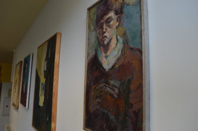 Obrazy powieszone na ścianie prezentujące archiwalne malarstwo uczniów ZSP. Na obrazach postaci oraz architektura w żywych i kontrastowych kolorach.