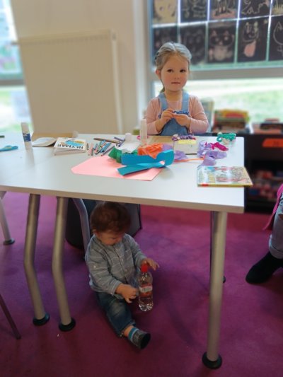 Dziewczynka robiąca wycinankę z bibuły. Chłopiec bawi się pod stołem.