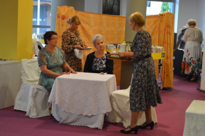 Dwie kobiety siedzą przy nakrytym białym stole a dwie kobiety im usługują - fragment spektaklu Dom Kobiet w wykonaniu grupy Teatru Baccalaureus.